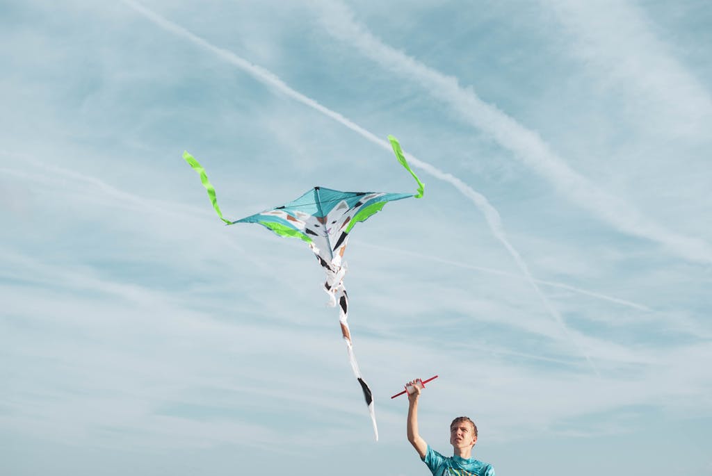 A young man in a blue shirt flying a colorful kite in a clear sky, enjoying the breeze.